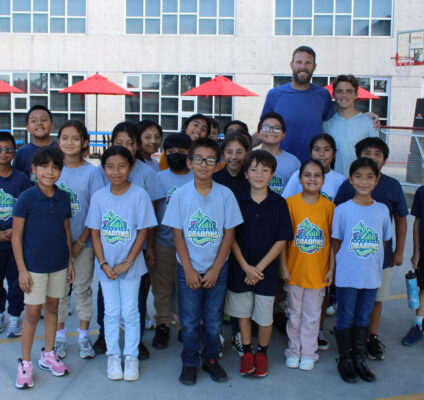 Chris Sale, a professional baseball pitcher, poses outdoors with a group of enthusiastic students from RCMA’s Immokalee Community Academy (ICA). The students, proudly wearing ICA-themed t-shirts and casual clothing, stand together smiling, with staff members and Sale in the back row. The vibrant scene takes place in front of the school building, with red patio umbrellas and a basketball hoop in the background, highlighting the engaging and welcoming environment of RCMA’s Immokalee Community Academy.