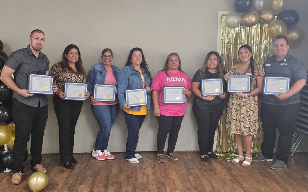 Group of RCMA Family Support Workers standing together indoors, holding certificates of completion for the Family Development Credential Program. The group is smiling, with a backdrop featuring gold and black balloons and a golden tinsel curtain.