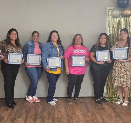 Group of RCMA Family Support Workers standing together indoors, holding certificates of completion for the Family Development Credential Program. The group is smiling, with a backdrop featuring gold and black balloons and a golden tinsel curtain.