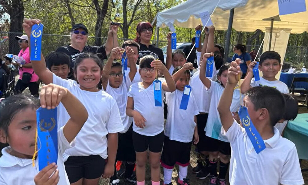 A joyful group of students from RCMA’s Wimauma Community Academy proudly holds up their blue first-place ribbons after participating in a soccer tournament during the school’s Fall Festival. The children, dressed in white shirts and black shorts, beam with pride as they pose outdoors under a sunny sky. Behind them, teachers and parents cheerfully join in the celebration, with tents and trees adding to the festive atmosphere.