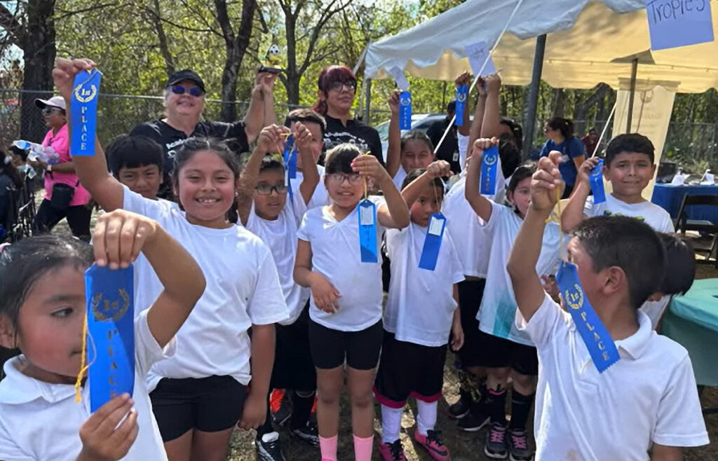 A joyful group of students from RCMA’s Wimauma Community Academy proudly holds up their blue first-place ribbons after participating in a soccer tournament during the school’s Fall Festival. The children, dressed in white shirts and black shorts, beam with pride as they pose outdoors under a sunny sky. Behind them, teachers and parents cheerfully join in the celebration, with tents and trees adding to the festive atmosphere.