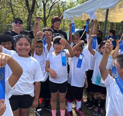 A joyful group of students from RCMA’s Wimauma Community Academy proudly holds up their blue first-place ribbons after participating in a soccer tournament during the school’s Fall Festival. The children, dressed in white shirts and black shorts, beam with pride as they pose outdoors under a sunny sky. Behind them, teachers and parents cheerfully join in the celebration, with tents and trees adding to the festive atmosphere.