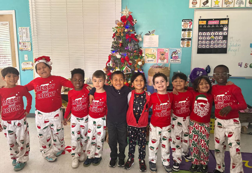 A joyful group of children from RCMA stands together in festive holiday attire, including matching red shirts that read 'Merry and Bright' and Santa-themed pajama pants. They smile brightly in front of a beautifully decorated Christmas tree adorned with ornaments, lights, and a shining star on top. The classroom background features cheerful wall decorations, educational posters, and a holiday calendar, creating a warm and festive atmosphere.