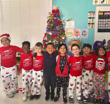 A joyful group of children from RCMA stands together in festive holiday attire, including matching red shirts that read 'Merry and Bright' and Santa-themed pajama pants. They smile brightly in front of a beautifully decorated Christmas tree adorned with ornaments, lights, and a shining star on top. The classroom background features cheerful wall decorations, educational posters, and a holiday calendar, creating a warm and festive atmosphere.