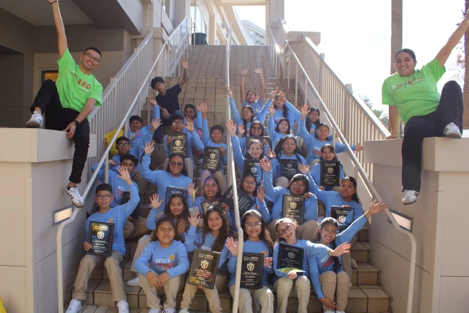 Immokalee Community Academy National Beta Club students and staff celebrating on outdoor stairs while holding awards and plaques.